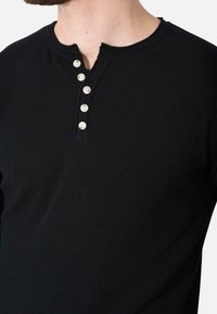 Maglietta henley a maniche lunghe nera in tessuto testurizzato, con patta a tre bottoni di colore chiaro e scollatura a girocollo.