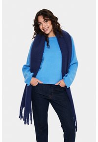 Pull bleu avec un ourlet et des poignets côtelés, associé à une écharpe à franges bleu foncé, portée sur un jean foncé. L'écharpe est douce et texturée.
