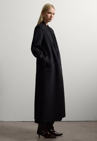 Lungo cappotto nero in lana con design doppiopetto, tasche laterali e silhouette strutturata, abbinato a stivali neri a punta.