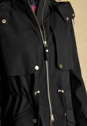 Schwarze Jacke mit goldfarbenen Beschlägen, Reißverschluss und Druckknopfverschlüssen, verstellbarem Kordelzug in der Taille und großen Fronttaschen; glatte Textur.