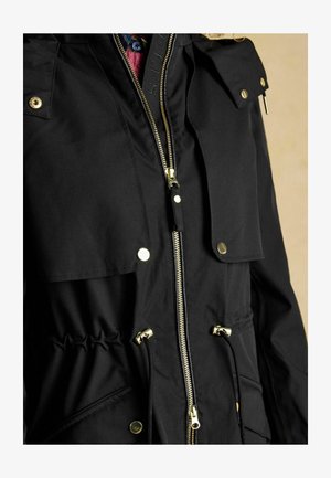 Schwarze Jacke mit goldfarbenen Beschlägen, Reißverschluss und Druckknopfverschlüssen, verstellbarem Kordelzug in der Taille und großen Fronttaschen; glatte Textur.