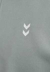 Grønt struktureret stof har et hvidt dobbelt-chevron logo på venstre side, hvilket tilføjer kontrast til materialets overflade.