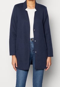 Cárdigan azul marino sin cuello, con mangas largas y dos bolsillos delanteros. Fabricado con una tela suave de textura lisa y cierres a presión.