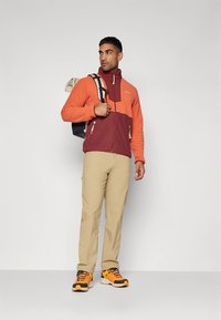 Chaqueta de forro polar con bloques de color naranja y burdeos, cierre de cremallera y bolsillos laterales; combinada con pantalones beige y zapatillas naranja y negras.