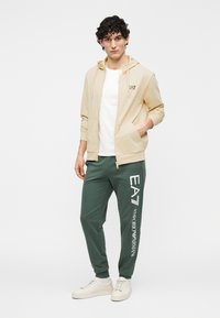 Lichtbeige capuchonvest met rits over een wit T-shirt, gecombineerd met groene joggers met witte logo's. Witte sneakers maken de outfit compleet.
