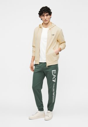 Veste à capuche beige clair zippée sur un t-shirt blanc, associée à un pantalon de jogging vert avec un logo blanc. Des baskets blanches complètent la tenue.