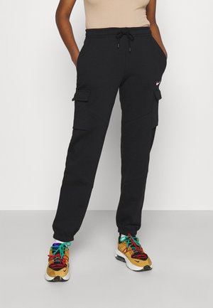 Personne portant un jogging cargo noir avec des poches latérales et des chaussures de sport multicolores, debout les mains dans les poches.
