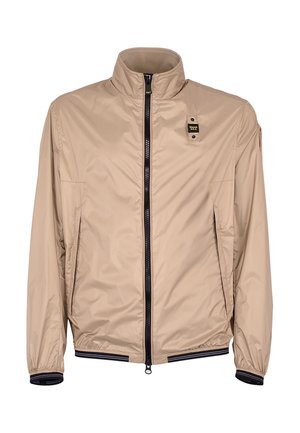 Beige lichte jas met opstaande kraag, zwarte ritssluiting aan de voorkant, elastische manchetten en zoom, en een klein zwart-geel logo op de borst.