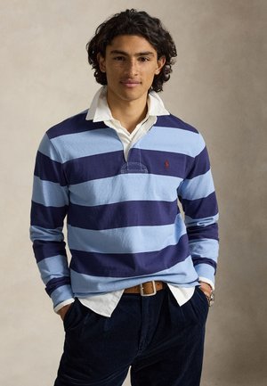 Jeune homme aux cheveux bouclés portant un maillot de rugby bleu rayé par-dessus une chemise blanche à col, les mains dans les poches, debout devant un fond neutre.