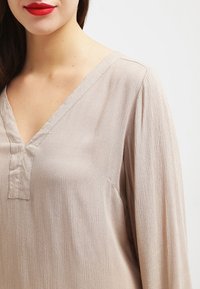 Blouse beige texturée avec un col en V et des manches longues. Dotée d'une patte de boutonnage ornée de boutons près du col. Tissu lisse et léger.