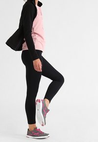 Persoon in zwarte leggings, roze shirt en zwarte jack, die één been omhoog til en grijze en roze sportschoenen laat zien, tegen een witte achtergrond.