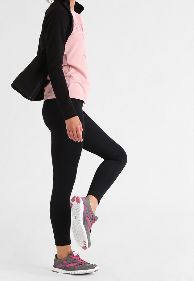 Persoon in zwarte leggings, roze shirt en zwarte jack, die één been omhoog til en grijze en roze sportschoenen laat zien, tegen een witte achtergrond.
