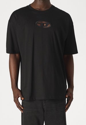 Person iført en løs sort t-shirt med en lille oval udskæring med et stiliseret "D" design på brystet, kombineret med mørke bukser.