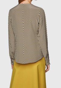 Blouse à motifs avec un design géométrique crème et marron, manches longues, col rond et tissu lisse, associée à une jupe jaune.