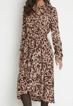 Vrouw draagt een midi-jurk met lange mouwen, knoopsluiting, een abstract patroon in bruin, beige en zwart en een geknoopte tailleceintuur.
