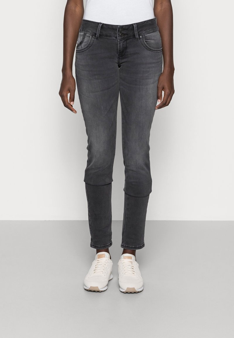 Personne portant un jean slim noir délavé avec fermeture double bouton et des baskets blanches, debout devant un fond uni.