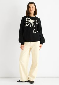Maglione nero lavorato a maglia con un grande design di fiocco bianco al centro. Abbinato a pantaloni beige a gamba larga e scarpe nere basse.