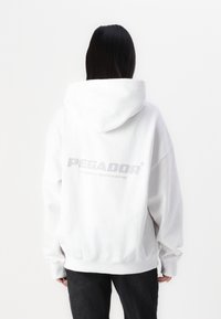 Hvit oversized hettegenser med en frontlomme og ribbede mansjetter. Baksiden viser logoen "PEGADOR" i stor grå tekst. Myk stofftekstur.