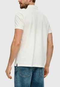 Weißes Polo-Shirt mit kurzen Ärmeln, klassischen Kragen, gerader Passform und seitlichen Schlitzen, getragen mit blauen Jeans.