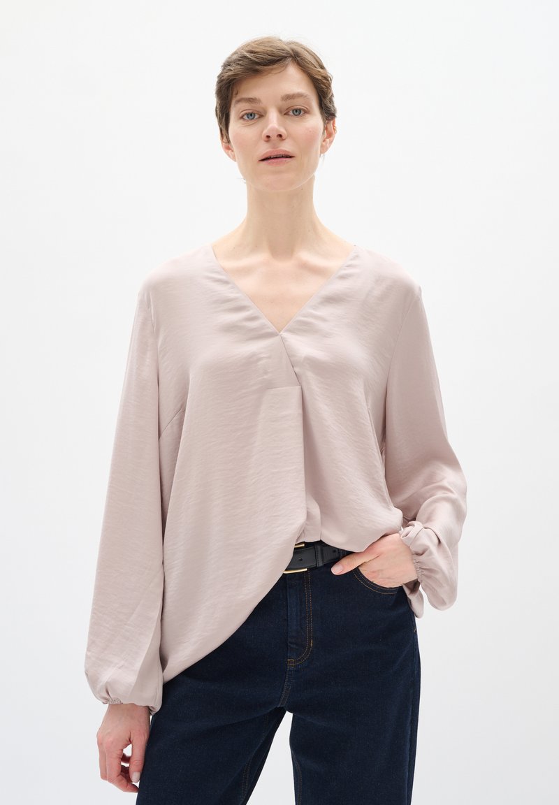 Blusa rosa chiaro con scollo a V, maniche larghe e polsini arricciati, realizzata in un tessuto morbido, abbinata a jeans di denim scuri e una cintura nera.