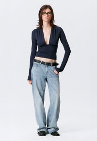 Top de manga larga azul marino con escote en V profundo y pliegues, combinado con pantalones rectos de mezclilla azul claros y un cinturón negro. Zapatos negros.