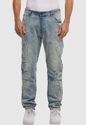 Mann trägt hellblaue, abgenutzte Jeans und weiße Sneakers, steht vor einem schlichten Hintergrund.