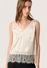 Top senza maniche color crema con scollatura a V; presenta un tessuto testurizzato e un bordo in pizzo con dettagli frangiati.