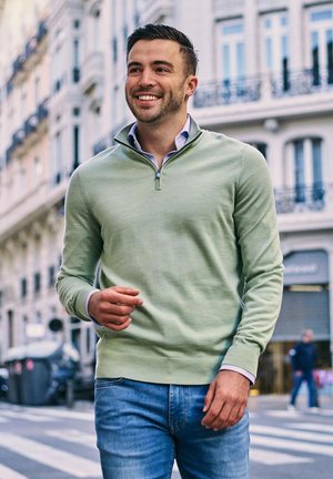 Lachende jonge man in groene trui en spijkerbroek loopt op een stadstraat met gebouwen in Europese stijl op de achtergrond.