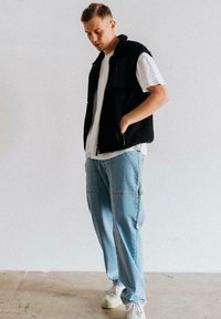 Schwarze Fleece-Weste über einem weißen Hemd, kombiniert mit hellblauen Cargo-Hosen. Das Outfit hat eine lockere Passform und minimale Hardware-Details.