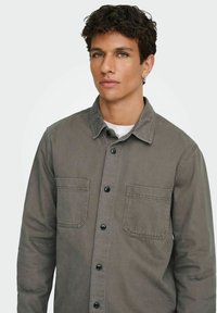 Camisa de algodón gris con dos bolsillos en el pecho, cierre con botones en la parte frontal, cuello puntiagudo y botones negros. La tela tiene una textura suave y mate.