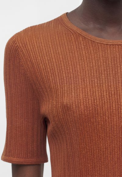 Κοντινό πλάνο ενός σκουρόχρωμου ατόμου που φοράει ένα σκουρόχρωμο, ριμπ πλεκτό κοντομάνικο τοπ με στρογγυλή λαιμόκοψη.