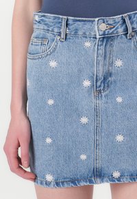 Light blue denim skirt with white embroidered flower patterns, five-pocket design, and a frayed hem. Front button closure and belt loops.