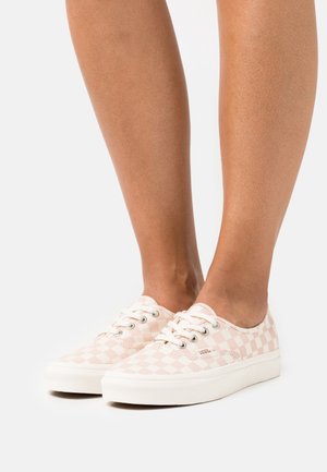 Piernas con zapatillas Vans bajas con patrón de cuadros rosa claro y blanco y cordones blancos sobre un fondo blanco.