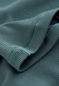 Tissu côtelé plié de couleur sarcelle avec coutures visibles et surpiqûres fines, mettant en valeur la texture et les détails du vêtement.