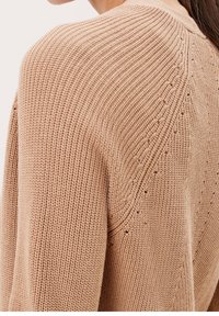 Stickad cardigan i beige, med ett texturerat mönster och öppna sömmar längs axelsömmarna samt en avslappnad passform.
