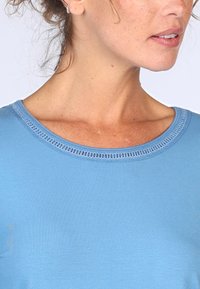 Helles blaues T-Shirt mit glatter Textur und Rundhalsausschnitt, der mit kunstvollen dekorativen Nähten entlang des Kragenrandes verziert ist.