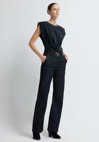 Námořnický bezrukávový top s texturovanou látkou, v kombinaci s tmavými džínovými kalhotami s vysokým pasem, které mají viditelné švy a kovovou sponu.