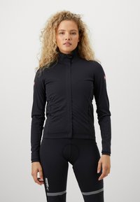 Frau mit lockigem, blonden Haar, die eine schwarze Zip-Jacke und schwarze Sportleggings mit reflektierenden Streifen trägt, steht vor einem schlichten weißen Hintergrund.