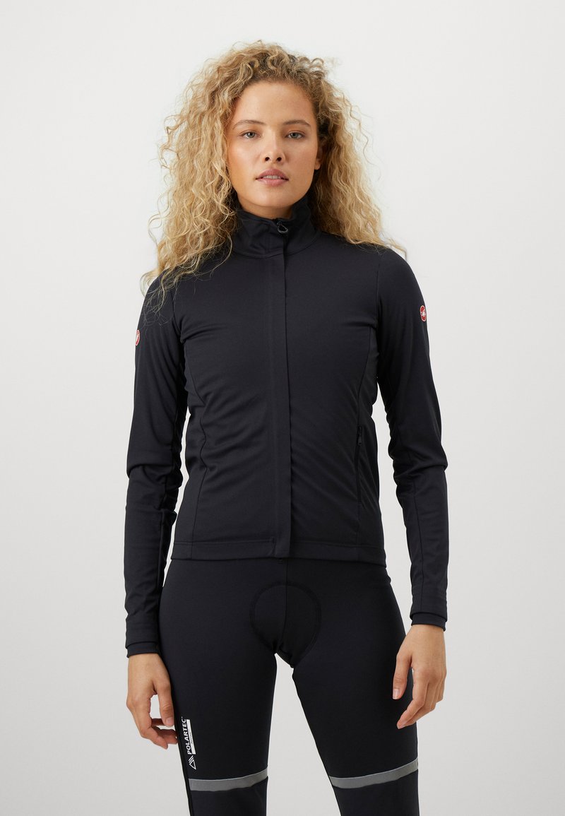Frau mit lockigem, blonden Haar, die eine schwarze Zip-Jacke und schwarze Sportleggings mit reflektierenden Streifen trägt, steht vor einem schlichten weißen Hintergrund.