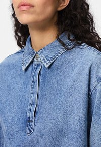 Jeansskjorta i ljusblå färg med knäppkrage, tryckknappslås och subtil textur. Tyget visar ett klassiskt jeansmönster.