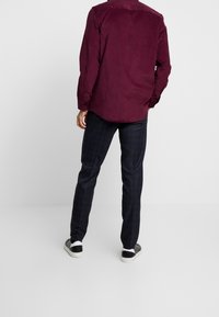 Langes Hemd aus rotem Cord mit gebogenem Saum, kombiniert mit dunkel karierten Hosen. Schwarze Slip-On-Schuhe vervollständigen das Outfit.