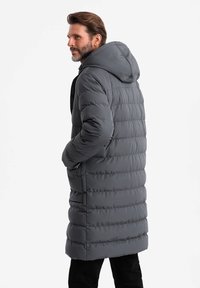 Cappotto trapuntato lungo e grigio con cappuccio, design trapuntato e tasche laterali. Il materiale sembra liscio e isolato per garantire calore.