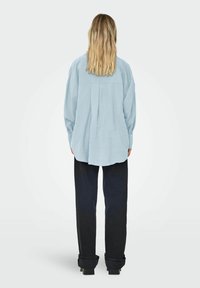 Camicia oversized azzurro chiaro con piega sul retro, maniche lunghe e polsini abbottonati, abbinata a pantaloni ampi blu scuro.