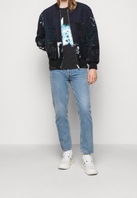 Bomberjakke i marineblå uldblanding med grønne abstrakte mønstre, lynlås, ribbede ærmer, parret med sort t-shirt og lyseblå jeans.