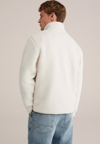 Giacca in pile bianca con colletto alto, caratterizzata da una texture morbida e una vestibilità rilassata. Abbinata a jeans azzurri chiari, con cuciture semplici.