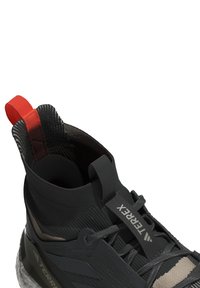 Chaussure de sport noire et beige avec un design montante, une tige texturée et une languette rouge. Équipée de lacets de marque et d'une semelle solide.