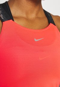 Débardeur de sport Nike en corail vif. Présente une texture lisse, des bretelles noires contrastantes et un logo argenté réfléchissant au centre.