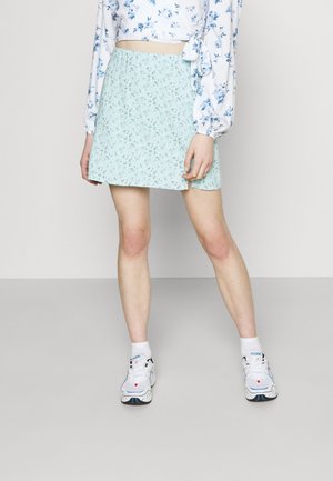 Osoba ubrana w jasno niebieską kwiecistą mini spódniczkę, biały kwiecisty długi crop top, białe skarpetki oraz białe sportowe trampki na jednolitym tle.