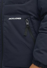 Tamsiai mėlynas pūkuotas švarkas, turintis užtrauktuko kišenę, elastingas rankogalius ir baltai atspaudintą logotipą. Lygus tekstūra su šiuolaikišku dizainu.