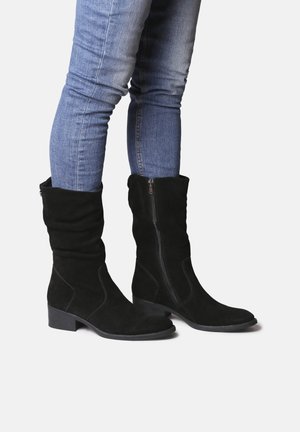 Bottes en suédine noires jusqu'au genou avec un design fluide, talon plat, fermeture éclair sur le côté et texture lisse, associées à un jean en denim bleu.